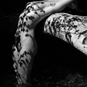 Cień od liści. Naga dziewczyna stojąca po kolana w wodzie, oparta o konar drzewa na którą padają cienie z gałęzi